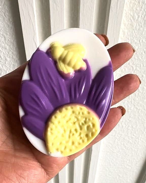 Jabón ovalado blanco con una flor morada, el centro amarillo con relieve y una abeja amarilla, sostenido en la mano.
