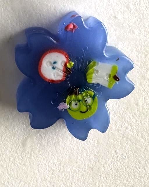 Jabón azul translúcido en forma de flor con tres figuritas de colores incrustadas en el centro.
