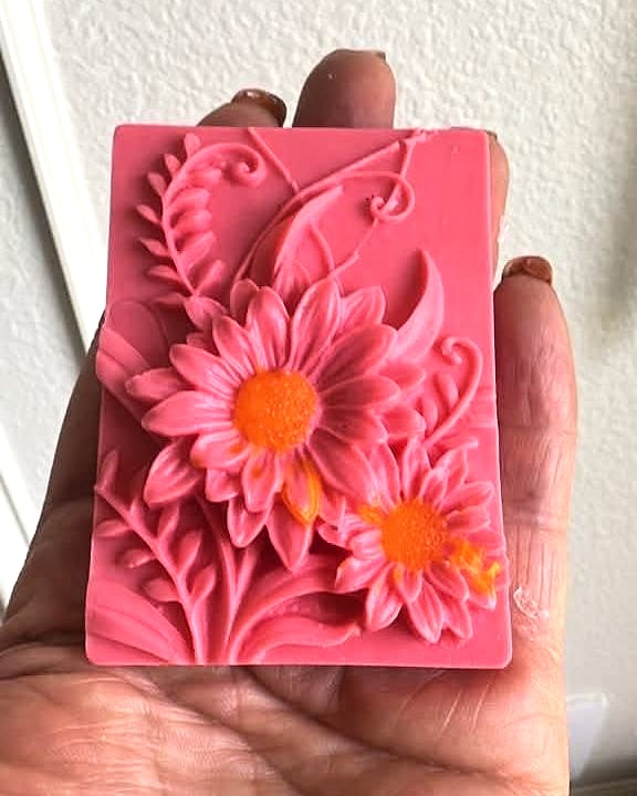 Jabón rectangular en relieve color coral con dos gerberas de centro naranja y hojas, sostenido en la mano.