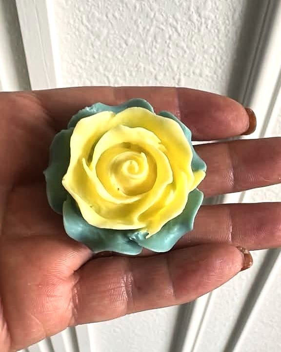 Jabón en forma de rosa amarilla sobre una base verde azulada, sostenido en la palma de una mano.